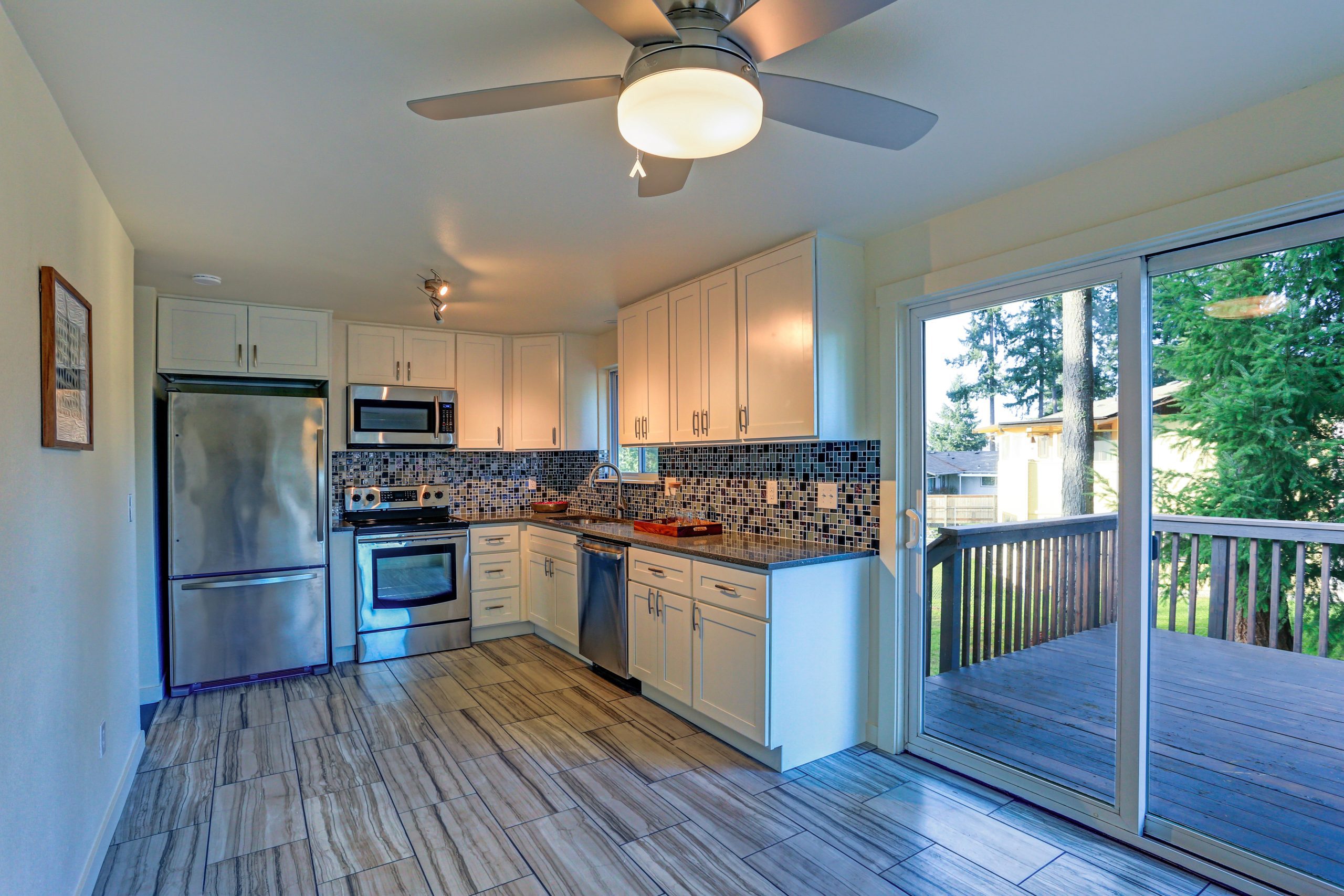L-shape kitchen room design with white cabinets, brown granite countertops, mosaic backsplash, built in stainless steel appliances and wide plank hardwood floor.