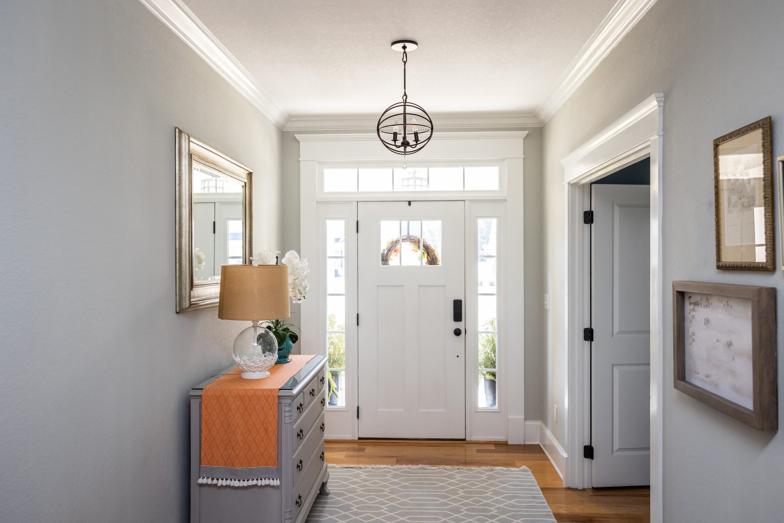 An open large and wide interior front door hallway foyer with transom, hanging light fixture, coastal colors and entry way table and wood floors.